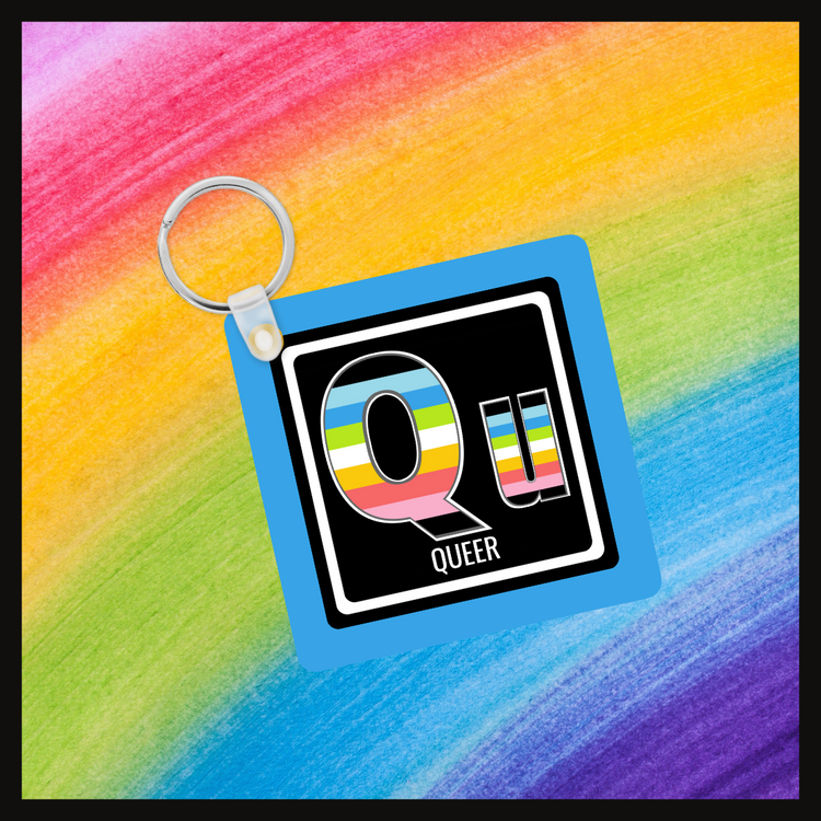 Keychain with the Element symbol (Qu) and name (Queer)with a design based on the colors of that element’s flag in a black square with a blue background. Behind the keychain is a rainbow background.