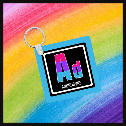 Keychain with the element symbol (Ad) and name (Androgyne) with a design based on the colors of that element’s flag in a black square with a blue background. Behind the keychain is a rainbow background.