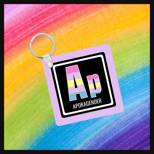 Keychain with the element symbol (Ap) and name (Aporagender) with a design based on the colors of that element’s flag in a black square with a lavender background. Behind the keychain is a rainbow background.