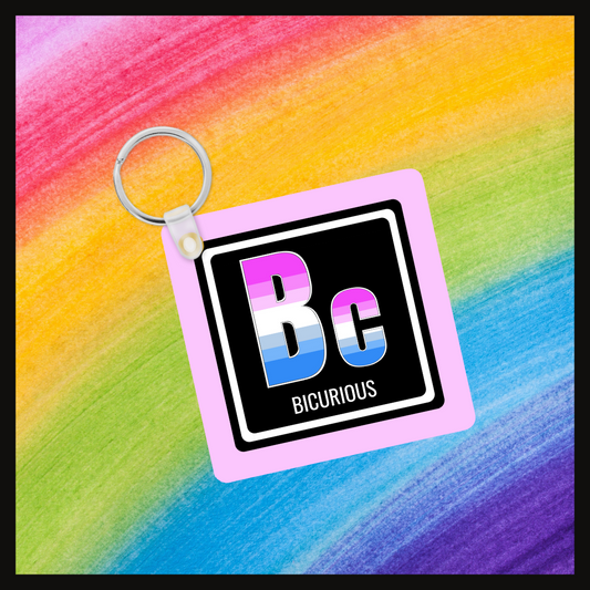 Keychain with the element symbol (Bc) and name (Bicurious) with a design based on the colors of that element’s flag in a black square with a pink background. Behind the keychain is a rainbow background.