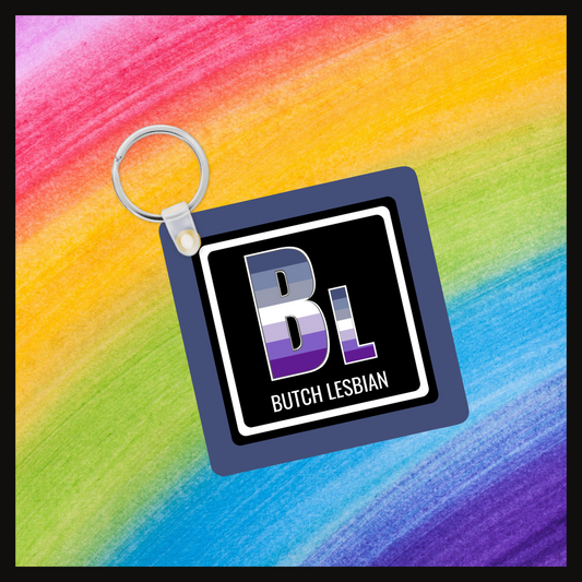 Keychain with the element symbol (Bl) and name (Butch Lesbian) with a design based on the colors of that element’s flag in a black square with a blue background. Behind the keychain is a rainbow background.