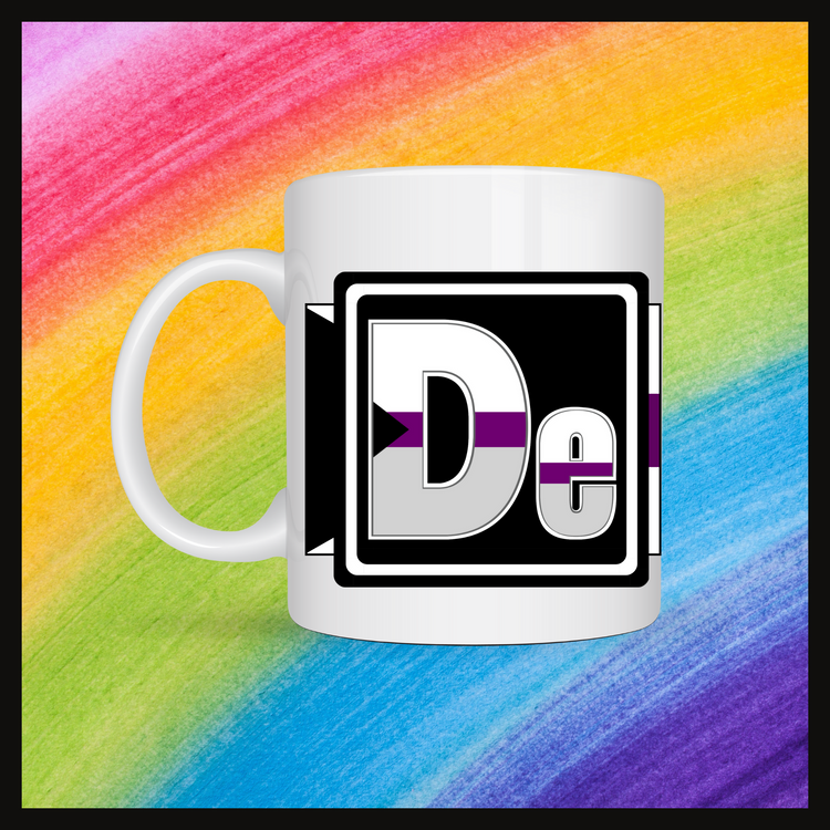 White mug with a 3 inch square containing the element’s symbol (De). Symbol is made with the colors of that element’s flag in a black square with the element’s flag behind the square. Behind the mug is a rainbow background.