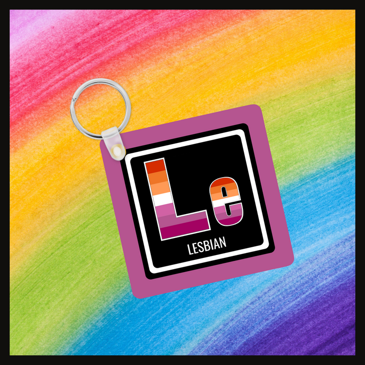 Keychain with the element symbol (Le) and name (Lesbian) with a design based on the colors of that element’s flag in a black square with a purple background. Behind the keychain is a rainbow background.