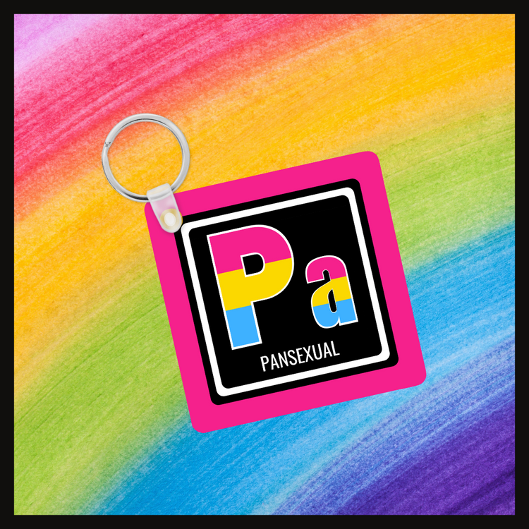Keychain with the element symbol (Pa) and name (Pansexual) with a design based on the colors of that element’s flag in a black square with a pink background. Behind the keychain is a rainbow background.
