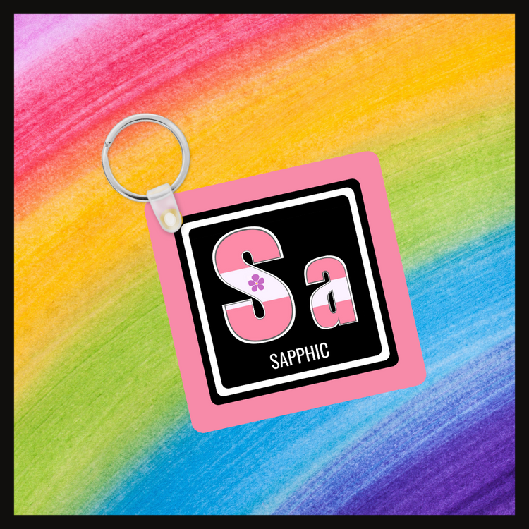 Keychain with the element symbol (Sa) and name (Sapphic) with a design based on the colors of that element’s flag in a black square with a pink background. Behind the keychain is a rainbow background.