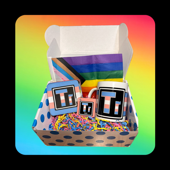 This is our Trans Element Gift Box - on a rainbow background inside a dotted blue box that holds a Queer Element Mug, Keychain and a set of our LGBTQ Coasters