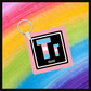 Keychain with the  Element symbol (Tr) and name (trans) with a design based on the colors of that element’s flag in a black square with a pink background. Behind the keychain is a rainbow background.