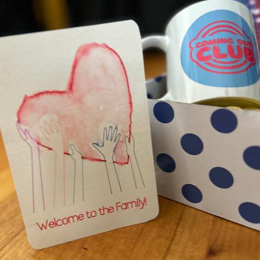 The contents of the box are inside a blue and white spotted box.  On the lef is the Welcome to The Family postcatd (with a watercolor red heart) Inside the box is a coming out club mug with blue/white background and red letters
