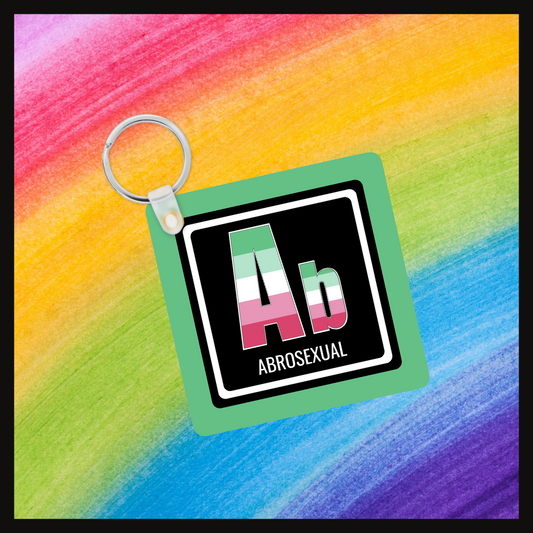 Keychain with the element symbol (Ab) and name (Abrosexual) with a design based on the colors of that element’s flag in a black square with a green background. Behind the keychain is a rainbow background.