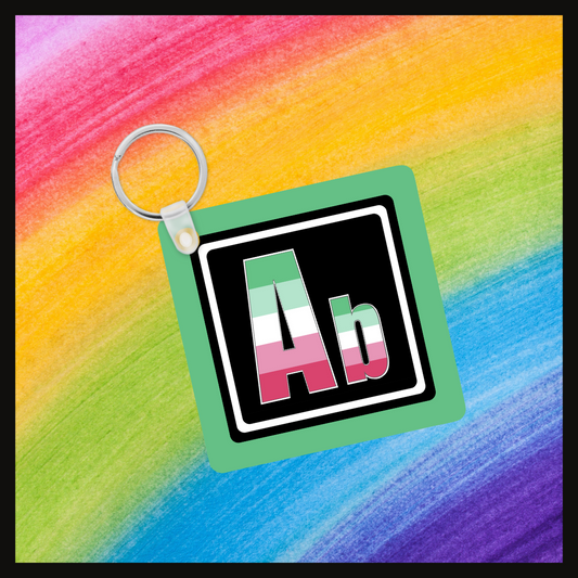 Keychain with the element symbol (Ab) with a design based on the colors of that element’s flag in a black square on a green background. Behind the keychain is a rainbow background.