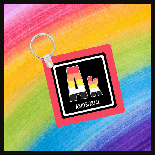 Keychain with the  Element symbol (Ak) and name (Akiosexual) with a design based on the colors of that element’s flag in a black square with a red background. Behind the keychain is a rainbow background.