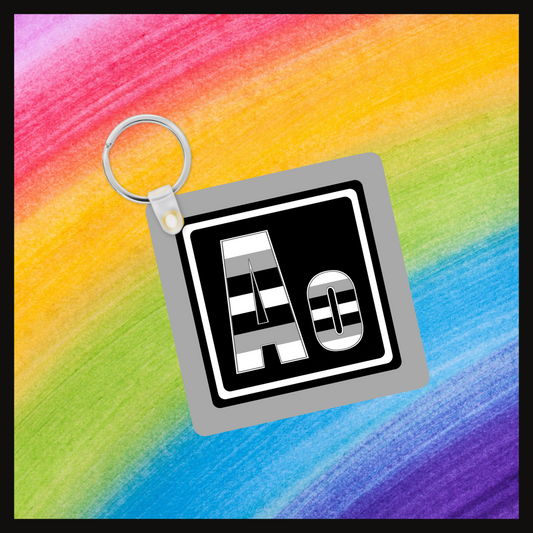 Keychain with the element symbol (Ao) with a design based on the colors of that element’s flag in a black square with a gray background. Behind the keychain is a rainbow background.