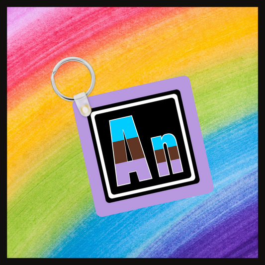  Keychain with the element symbol (An) with a design based on the colors of that element’s flag in a black square with a purple background. Behind the keychain is a rainbow background.