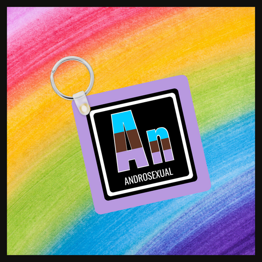  Keychain with the element symbol (An) and name (Androsexual) with a design based on the colors of that element’s flag in a black square with a purple background. Behind the keychain is a rainbow background.