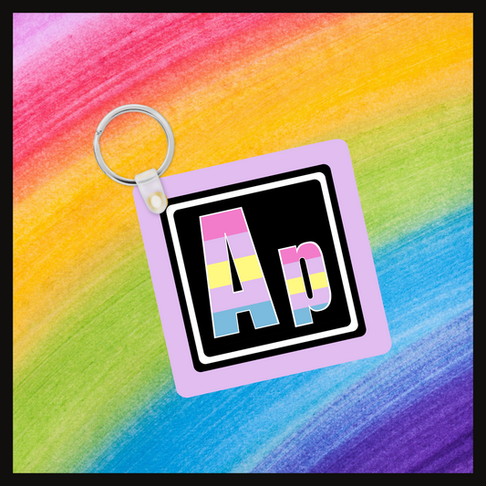 Keychain with the element symbol (Ap) with a design based on the colors of that element’s flag in a black square with a lavender background. Behind the keychain is a rainbow background.