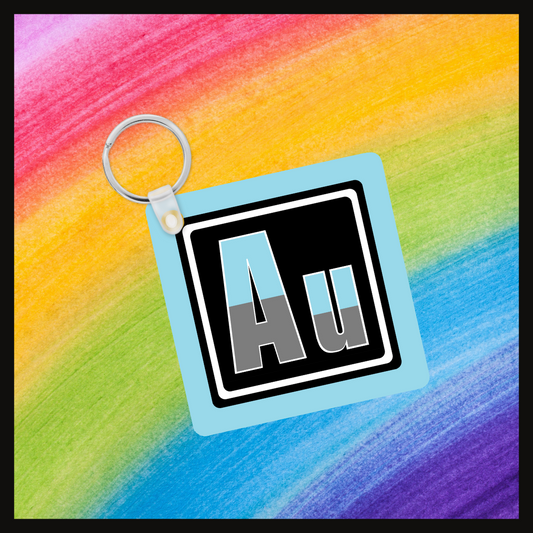 Keychain with the element symbol (Au) with a design based on the colors of that element’s flag in a black square with a blue background. Behind the keychain is a rainbow background.