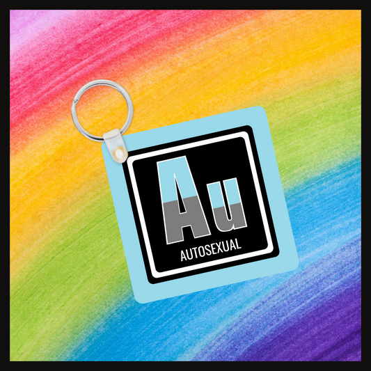 Keychain with the element symbol (Au) and name (Autosexual) with a design based on the colors of that element’s flag in a black square with a blue background. Behind the keychain is a rainbow background.