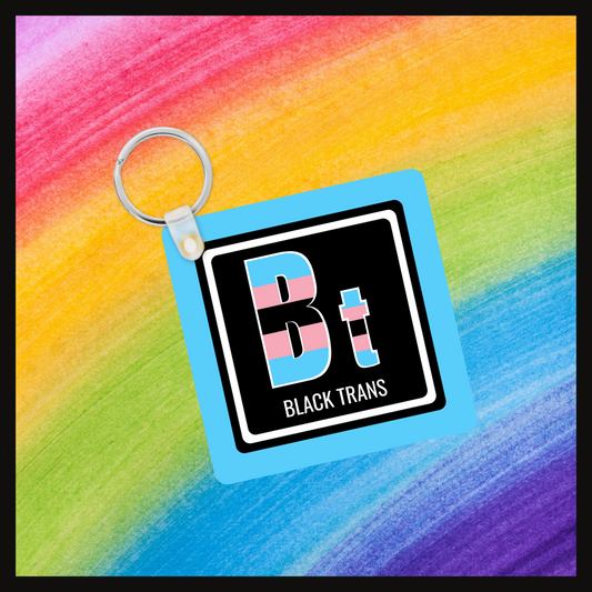 Keychain with the element symbol (Bt) and name (BlackTrans) with a design based on the colors of that element’s flag in a black square with a blue background. Behind the keychain is a rainbow background.