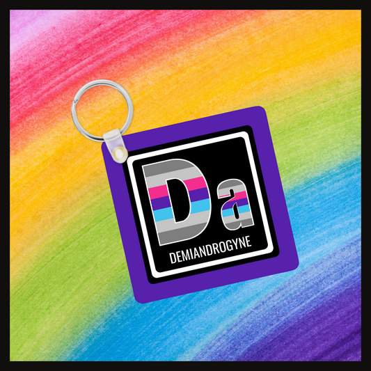 Keychain with the element symbol (Da) and name (Demiandrogyne) with a design based on the colors of that element’s flag in a black square on a purple background. Behind the keychain is a rainbow background.