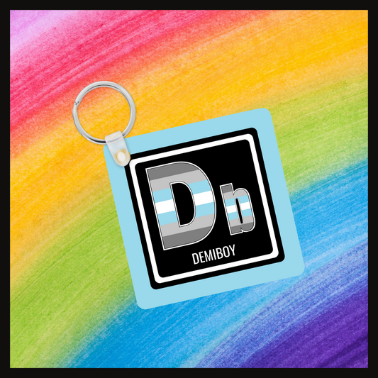 Keychain with the element symbol (Db) and name (demiboy) with a design based on the colors of that element’s flag in a black square with a blue background. Behind the keychain is a rainbow background.
