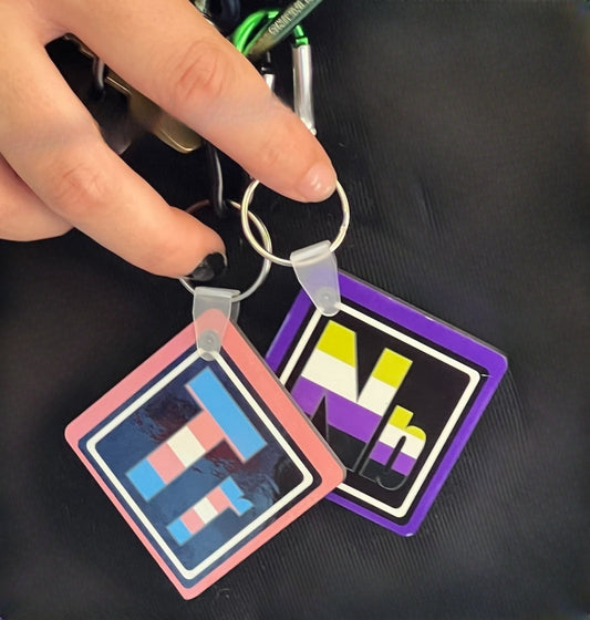 Two keychains being held by our model’s hands, showcasing our Trans and Non-Binary keychains. These keychains have the elements’ symbols (Tr & Nb) made with the colors of these element’s flags on a black background.